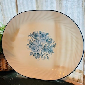 Puede incluir: Plato ovalado para servir con una superficie color crema y un diseño floral azul. El plato tiene un borde azul oscuro y un patrón de remolino alrededor del borde. El diseño floral presenta rosas azules y otras flores.