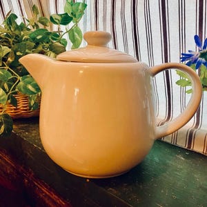 May include: A beige ceramic teapot with a lid and a curved handle. The teapot is sitting on a green surface, with a striped curtain and greenery in the background. The teapot is a light, neutral color.