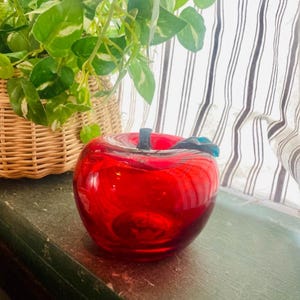 Puede incluir: Una vibrante escultura de manzana de cristal rojo con un tallo negro y un detalle de hoja azul. La pieza decorativa está colocada sobre una superficie verde, con una cesta de mimbre y una planta verde frondosa en el fondo.