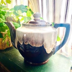 May include: A ceramic teapot in shades of brown, white, and blue. The teapot features a rounded body, a lid, and a curved spout. The handle is blue, and the base is brown. The teapot is displayed on a green surface.