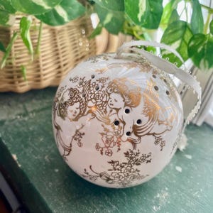 Może przedstawiać: Biała ceramiczna ozdoba w kształcie kuli ze złotymi aniołami i kwiatowymi wzorami. Ozdoba ma małe otwory i białą wstążkę do zawieszenia. Kula znajduje się na zielonej powierzchni, z roślinami i wiklinowym koszem w tle.