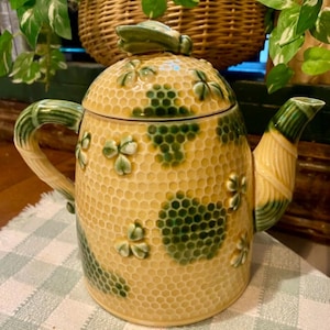 Puede incluir: Una tetera de cerámica con un patrón de panal amarillo y detalles de trébol verde. La tetera tiene un asa y un pico curvos, y un asa de tapa en forma de hoja verde. La tetera está sobre un mantel a cuadros.