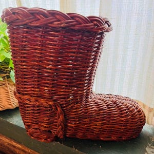 May include: A woven wicker boot-shaped basket in a rich reddish-brown hue. The basket features a braided trim around the top and a decorative braided detail on the side. The boot basket is designed for storage or decoration.
