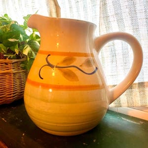 May include: A ceramic pitcher with a wide base and a curved handle. The pitcher is painted with horizontal yellow and green stripes, orange bands, and floral accents in blue and green. The pitcher is sitting on a green surface.