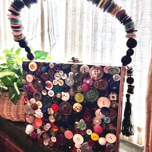 May include: A distinctive box handbag adorned with an assortment of colourful buttons. The handle is crafted from buttons and beads. A black tassel dangles from the side. This purse is a creative and striking accessory.