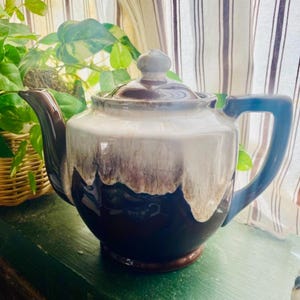 Peut inclure: Une théière en céramique émaillée brune et blanche avec une poignée bleue. La théière a un glaçage de style goutte à goutte avec des stries brunes et blanches.