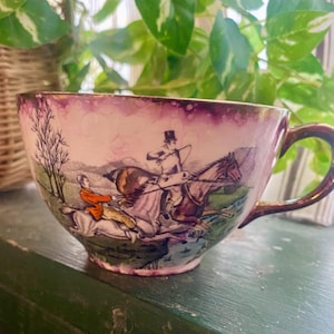 May include: A vintage teacup with a pink and purple glaze. The cup features a hand-painted scene of a man on horseback riding through a forest.