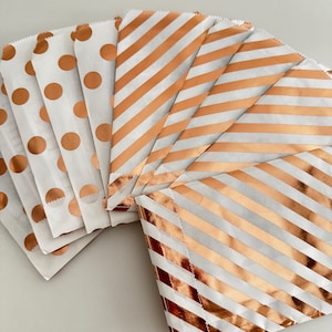 Könnte beinhalten: Eine Sammlung weißer Papiertüten mit kupferfarbenen Punkten und Streifen. Die Tüten sind fächerförmig angeordnet und zeigen die verschiedenen Muster. Die Tüten sind wahrscheinlich für Partygeschenke oder kleine Geschenke gedacht.