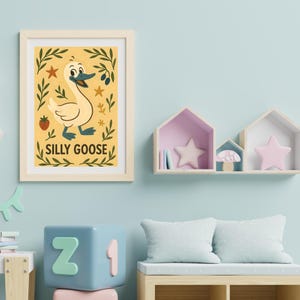 May include: A framed print featuring a whimsical illustration of a goose with the text "SILLY GOOSE". The artwork has a yellow background with decorative leaves, stars, and a strawberry. The frame is a light wood color.