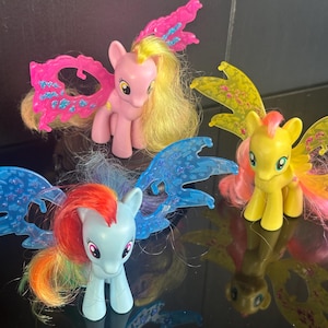 Puede incluir: Tres ponis de juguete coloridos con alas. Uno es azul claro con una melena arcoíris, otro es rosa con una melena amarilla y el tercero es amarillo con una melena rosa. Las alas son de varios colores con detalles brillantes.