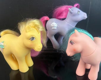 May include: Three vintage toy ponies in pastel colors. One is yellow with a pink and yellow mane, another is lavender with a pink mane, and the third is pink with a teal mane. Each pony has a unique design on its flank.