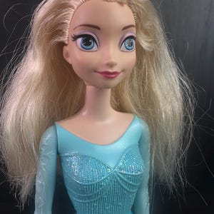 May include: A doll with blonde hair and blue eyes, wearing a light blue bodysuit with a sparkly bodice and long sleeves. The doll has a smiling expression and is a toy.