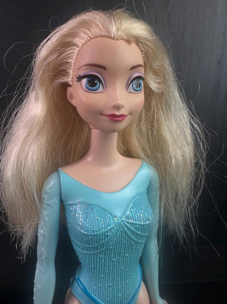 May include: A doll with blonde hair and blue eyes, wearing a light blue bodysuit with a sparkly bodice and long sleeves. The doll has a smiling expression and is a toy.