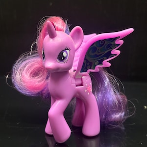 Op de afbeelding: Een paars speelgoedpony met een roze en paarse manen en staart. De pony heeft een hoorn en vleugels met een blauw en groen ontwerp. De pony heeft een sterontwerp op zijn flank.