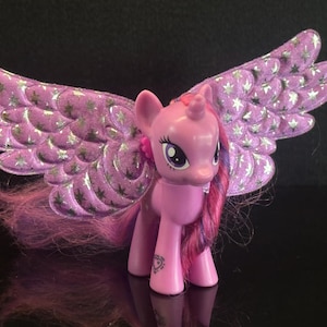 Op de afbeelding: Een roze plastic ponyfiguur met grote, glinsterende paarse vleugels versierd met een zilveren sterrenpatroon. De pony heeft lange, golvende manen met roze en paarse strepen en een klein hartvormig symbool op het voorbeen.