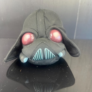 May include: A plush toy shaped like Darth Vader's helmet. The black fabric toy has large, red eyes and a stylized mouth with teal accents. The toy has ear flaps and is sitting on a reflective surface.