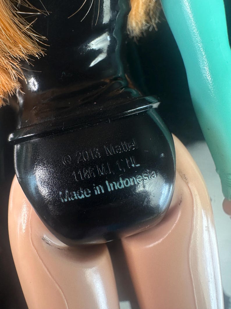 May include: Close-up of a doll's backside, featuring a black swimsuit bottom. The doll's skin is a light tan color. The text "&copy; 2013 Mattel 1186 MJ, 1, NL Made in Indonesia" is printed on the swimsuit bottom.