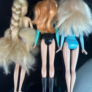May include: Three dolls with blonde, red, and blonde hair, each wearing a different outfit. The dolls are standing with their backs facing the viewer. The doll on the left has a long braid. The middle doll has red hair and black boots. The doll on the right has blonde hair and a blue outfit.