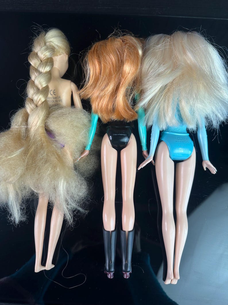 May include: Three dolls with blonde, red, and blonde hair, each wearing a different outfit. The dolls are standing with their backs facing the viewer. The doll on the left has a long braid. The middle doll has red hair and black boots. The doll on the right has blonde hair and a blue outfit.