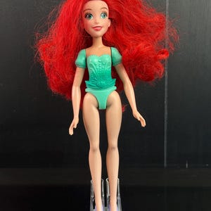 May include: A doll with long red hair, wearing a teal swimsuit and matching shoes. The doll has a fair complexion, green eyes, and a small pink bow in her hair. The doll is standing on a clear plastic stand.