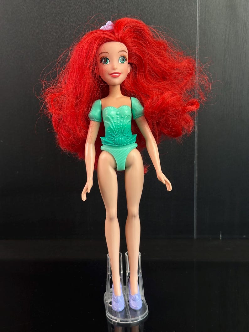 May include: A doll with long red hair, wearing a teal swimsuit and matching shoes. The doll has a fair complexion, green eyes, and a small pink bow in her hair. The doll is standing on a clear plastic stand.