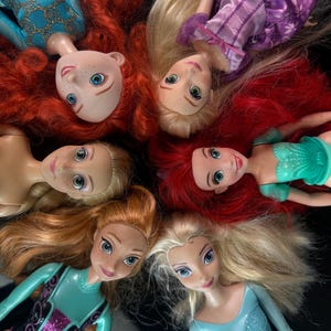 May include: A collection of six dolls with long hair and detailed facial features. The dolls are arranged in a circular pattern, with their heads facing inward. The dolls are wearing various outfits in shades of blue, green, and purple.