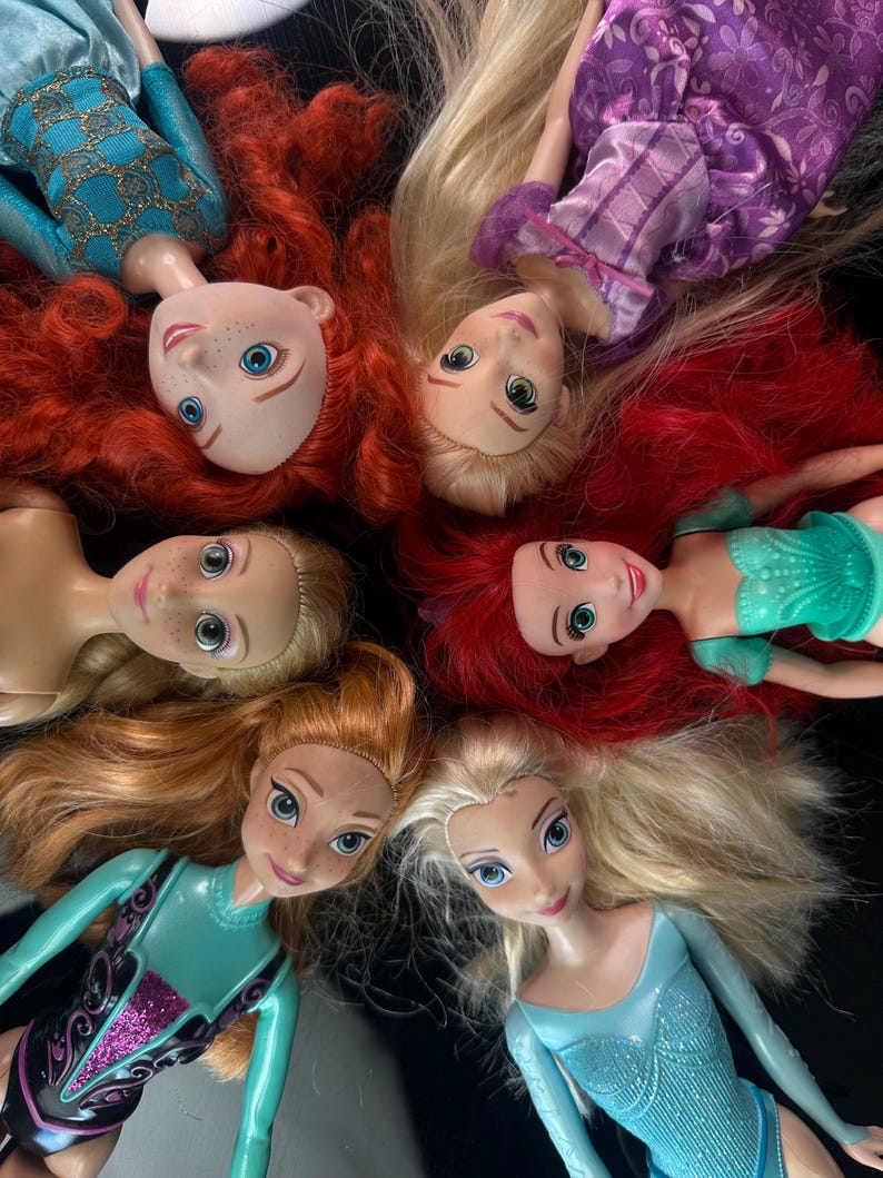 May include: A collection of six dolls with long hair and detailed facial features. The dolls are arranged in a circular pattern, with their heads facing inward. The dolls are wearing various outfits in shades of blue, green, and purple.