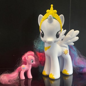 Puede incluir: Una figura de poni unicornio blanco con corona amarilla, alas y crin y cola multicolor, junto a una figura de poni rosa más pequeña con crin y cola rosas. El poni blanco presenta un símbolo de sol amarillo en el flanco y detalles amarillos.
