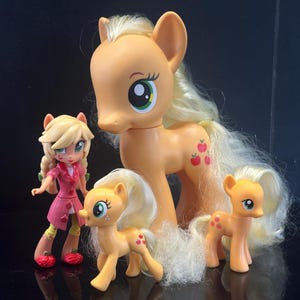 Puede incluir: Una colección de cuatro figuras de My Little Pony que incluye un poni naranja grande con crin rubia, dos ponis naranjas más pequeños con crines rubias y una muñeca de aspecto humano con cabello rubio que viste un abrigo rosa y zapatos rojos.