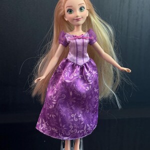 May include: A doll with long blonde hair and green eyes, wearing a purple dress with a floral pattern. The dress has a bodice with a pink bow and short purple sleeves. The doll is standing on a clear plastic stand.