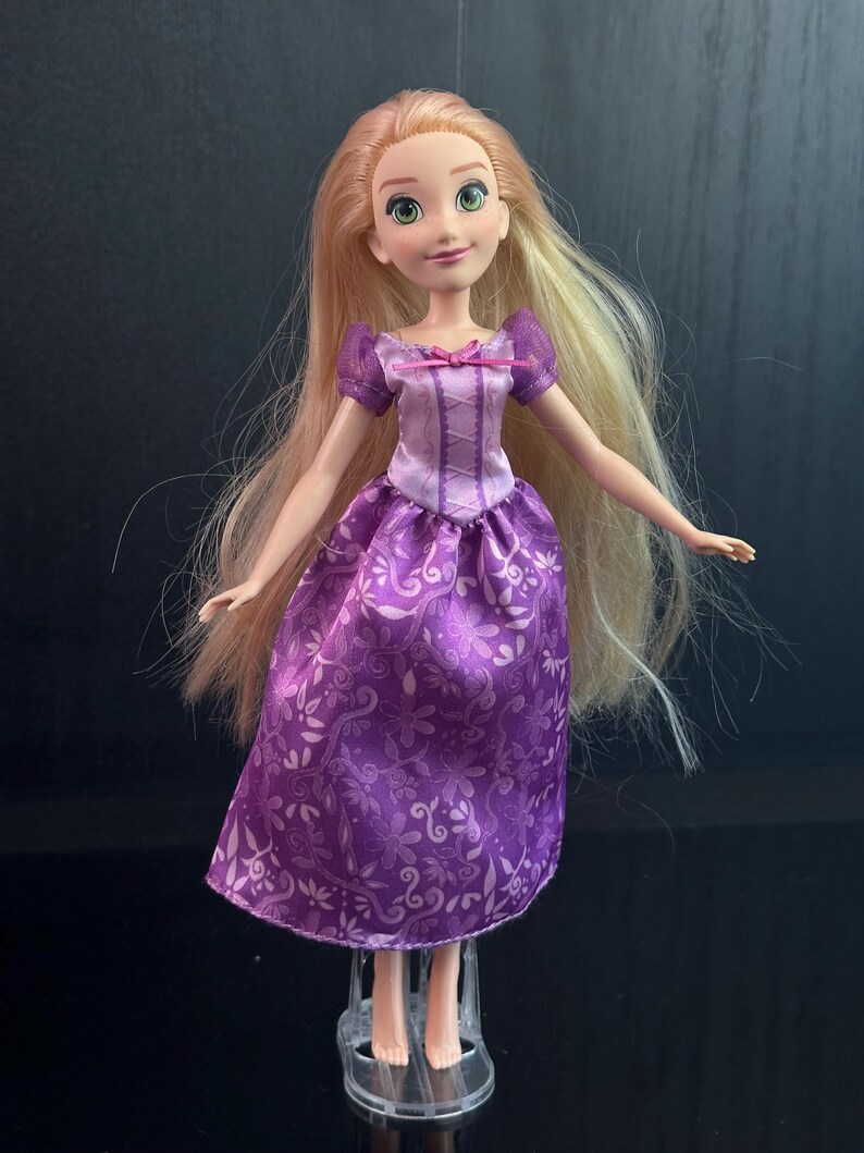 May include: A doll with long blonde hair and green eyes, wearing a purple dress with a floral pattern. The dress has a bodice with a pink bow and short purple sleeves. The doll is standing on a clear plastic stand.