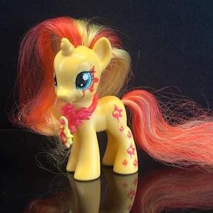 Puede incluir: Una figura de poni amarilla con un cuerno y una larga melena fluida de color rojo y amarillo. El poni tiene grandes ojos azules, un collar floral rojo con un colgante en forma de sol y marcas solares rojas en la mejilla y la pata trasera.