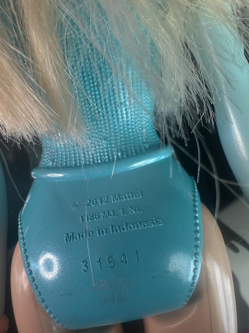 May include: Close-up of a doll's back, featuring a light blue swimsuit and blonde hair. The text on the doll reads "&copy; 2012 Mattel, Made in Indonesia" and the number "31541".