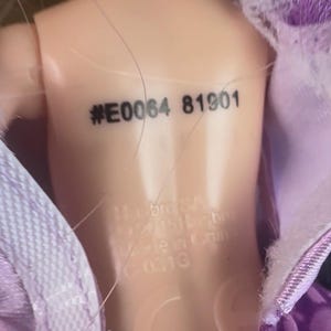 May include: Close-up of a doll's back with the stamped text "#E0064 81901". The doll's skin is a light peach color. The doll is partially covered by a purple fabric with a lace trim.