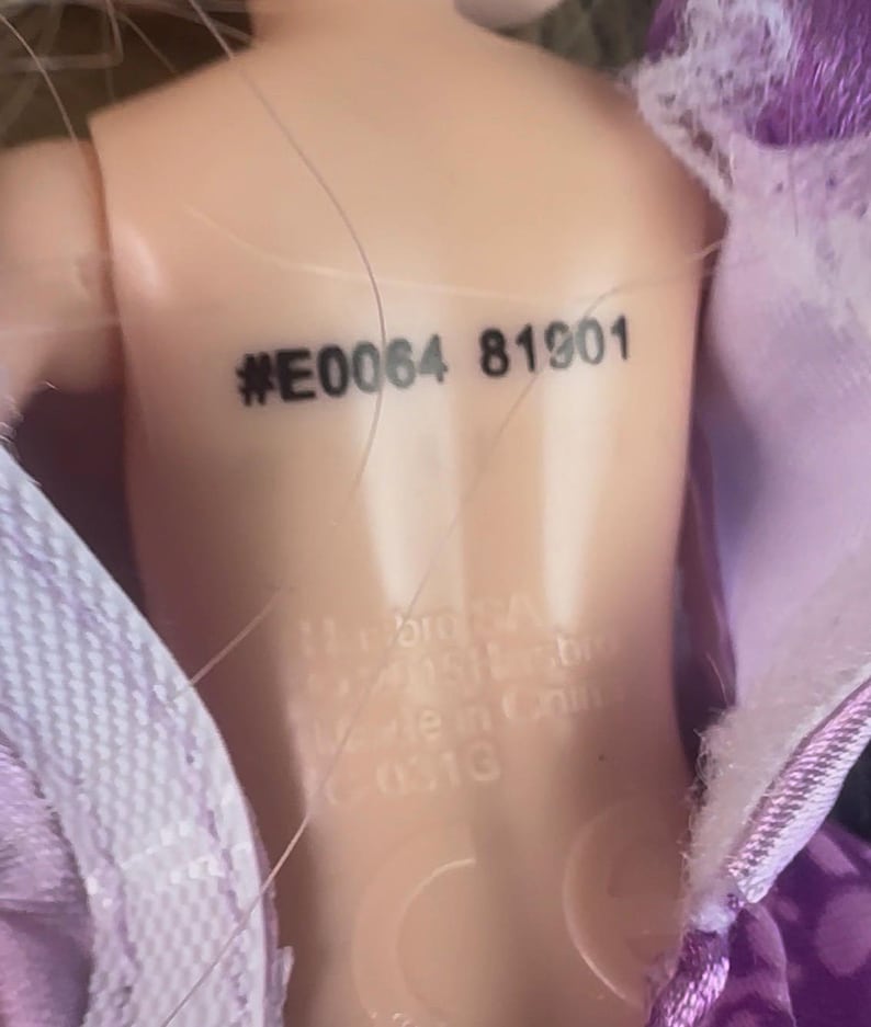 May include: Close-up of a doll's back with the stamped text "#E0064 81901". The doll's skin is a light peach color. The doll is partially covered by a purple fabric with a lace trim.