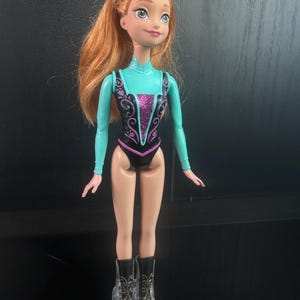 May include: A doll featuring red hair and a teal and black leotard with a pink center design. The doll has clear boots with black accents. The doll's arms and legs are a light skin tone. The doll is standing upright.