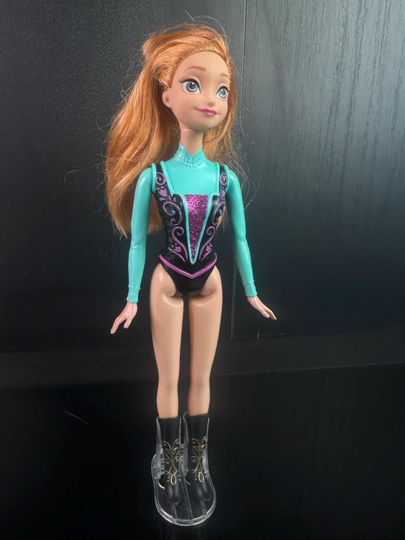 May include: A doll featuring red hair and a teal and black leotard with a pink center design. The doll has clear boots with black accents. The doll's arms and legs are a light skin tone. The doll is standing upright.