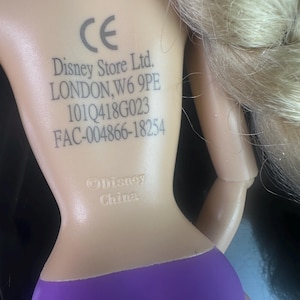 May include: A doll with blonde hair in a braid, wearing a purple swimsuit. The doll's back has printed text including "Disney Store Ltd. LONDON, W6 9PE" and a "CE" mark.