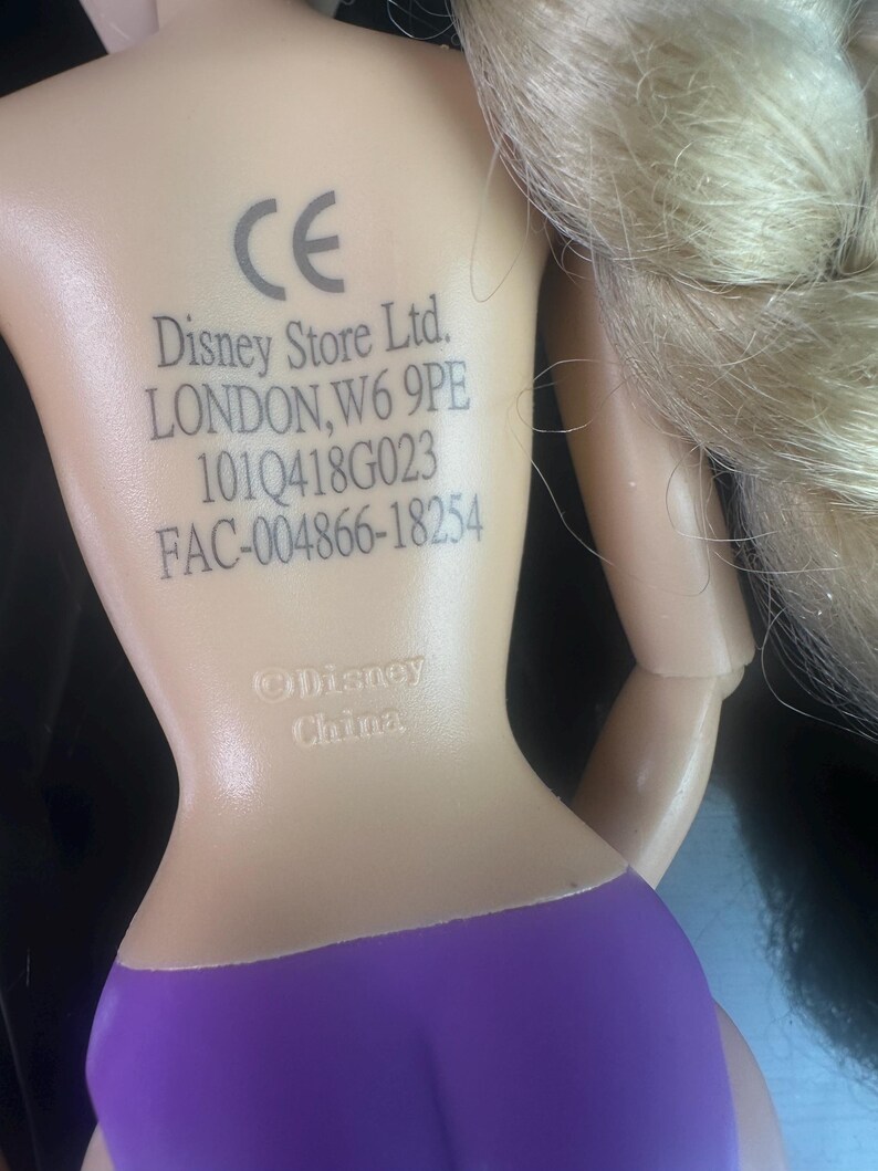 May include: A doll with blonde hair in a braid, wearing a purple swimsuit. The doll's back has printed text including "Disney Store Ltd. LONDON, W6 9PE" and a "CE" mark.