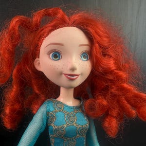 May include: A doll with fair skin, freckles, and bright blue eyes. The doll has vibrant, curly red hair and is wearing a teal dress with gold accents. The doll's expression is a slight smile. The doll is a character from a popular animated film.