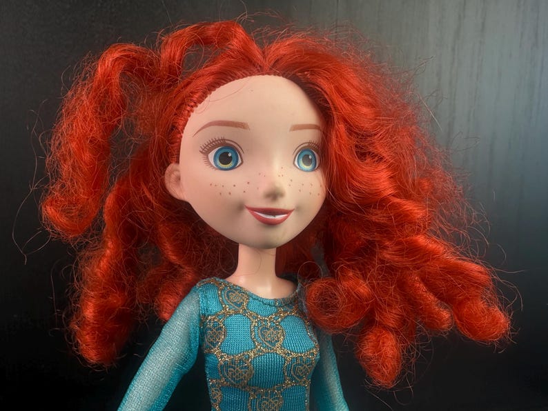May include: A doll with fair skin, freckles, and bright blue eyes. The doll has vibrant, curly red hair and is wearing a teal dress with gold accents. The doll's expression is a slight smile. The doll is a character from a popular animated film.