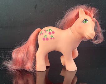 Op de afbeelding: Een perzikkleurige plastic speelgoedpony met lange, golvende roze manen en een staart. De pony heeft heldergroene ogen en een symbool op de flank bestaande uit vijf kleine, roze kerspictogrammen met groene steeltjes in een clusterpatroon.