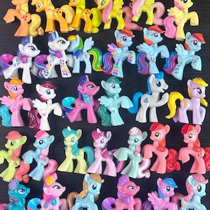 Op de afbeelding: Een verzameling van tweeëndertig kleine plastic ponyfiguurtjes gerangschikt in vijf rijen. Elke pony heeft een uniek kleurenschema, waaronder tinten roze, blauw, geel, wit en oranje, met gevormde manen, staarten en specifieke symbolen op de flanken.