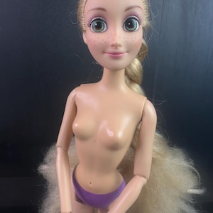 May include: A doll with blonde hair styled in a braid. The doll has green eyes, freckles, and is wearing purple underwear. The doll's arms and legs are articulated, and the doll is nude from the waist up.