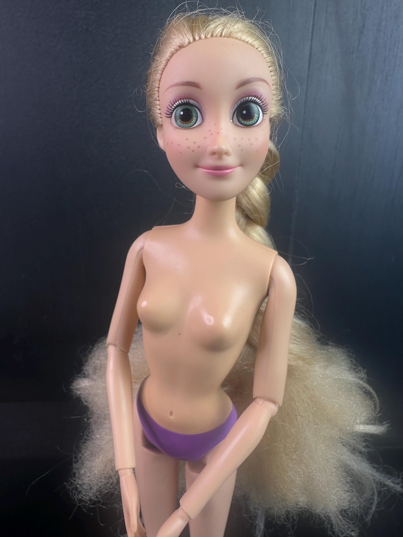 May include: A doll with blonde hair styled in a braid. The doll has green eyes, freckles, and is wearing purple underwear. The doll's arms and legs are articulated, and the doll is nude from the waist up.