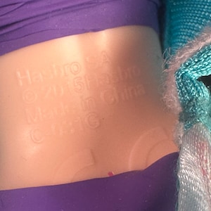 May include: Close-up of a doll's torso with a purple top and a light-colored plastic midsection. The text "Hasbro SA &copy; 2015 Hasbro Made in China C-031G" is embossed on the plastic.