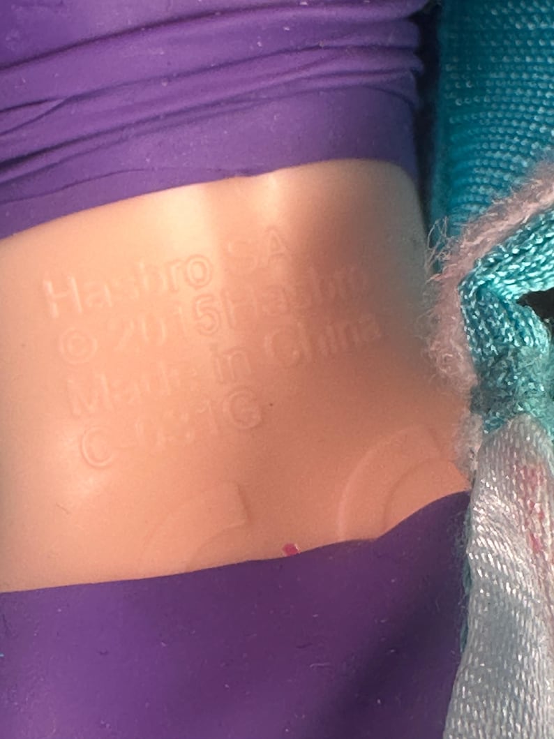 May include: Close-up of a doll's torso with a purple top and a light-colored plastic midsection. The text "Hasbro SA &copy; 2015 Hasbro Made in China C-031G" is embossed on the plastic.