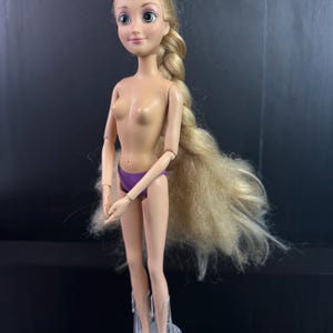 May include: A doll with long blonde hair in a braid, green eyes, and a purple bottom. The doll is standing on a clear plastic stand. The doll's arms and legs are articulated.