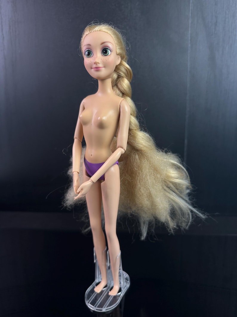 May include: A doll with long blonde hair in a braid, green eyes, and a purple bottom. The doll is standing on a clear plastic stand. The doll's arms and legs are articulated.