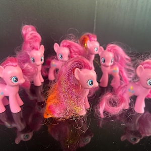 Può includere: Una collezione di pony giocattolo rosa con lunghi capelli rosa fluenti. Ogni pony ha occhi blu e un piccolo disegno colorato sul fianco. I pony sono disposti su una superficie riflettente.